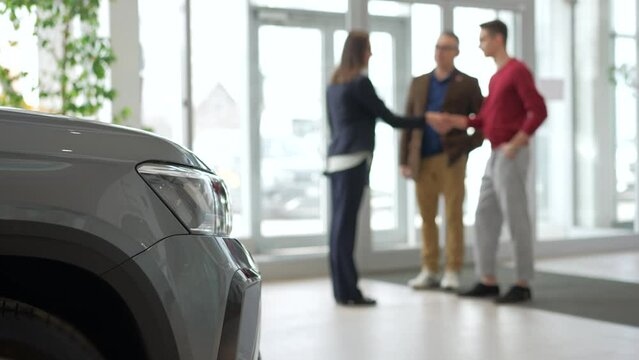 Two men are shaking hands with agent and talking while standing in car company spbd. 4k Blurred view of young son, elderly father make handshake with woman manager and talk with smiles, start choosing