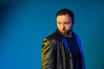Obraz premium a man looks over his shoulder to the side in the studio in a black leather coat. blue background
