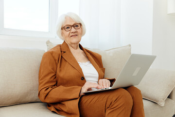 a cheerful elderly woman in a brown suit maintains a modern lifestyle using a laptop while sitting on the couch at home