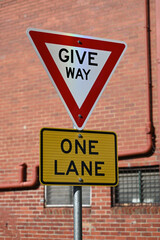 A red and white give way sign, featuring black text, above a yellow and black one lane sign, with a red brick building in the background