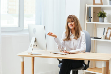 Smiling cheerful adorable blonde businesswoman worker freelancer look at camera raise hand up in light modern office. Happy employee work on computer online, enjoy successful business. Copy space