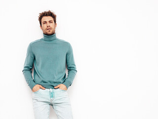 Fototapeta premium Portrait of handsome confident model. Sexy stylish man dressed in blue sweater and jeans. Fashion hipster male with curly hairstyle posing near white wall in studio. Isolated