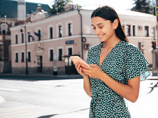 Young beautiful smiling hipster woman. Sexy carefree model posing in the street. Positive brunette female holding smartphone. Looking at cellphone screen. Using phone apps. Outdoors at sunny day