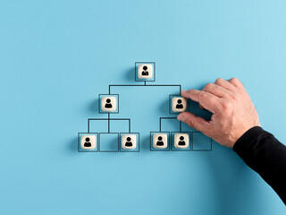 Male hand arranges company hierarchical organizational chart of wooden cubes on blue background. - obrazy, fototapety, plakaty
