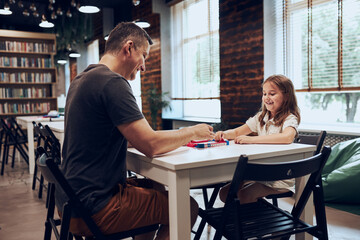 Father playing board game with her daughter in public library. Child spending time with dad after classes at primary school. Positive competition