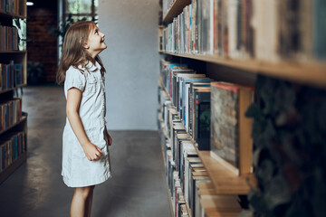 Schoolgirl looking at books on top bookshelf in school library. Smart girl selecting literature for reading. Benefits of everyday reading. Child curiosity. Back to school