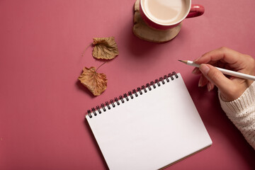 Table desk with notepad and female hand wit pencil and fall leaves on burgundy colored background. Autumn flat lay composition