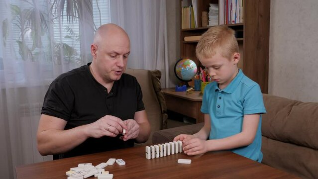 Cheerful father and son play with domino bones sitting at a wooden table, they put dominoes in a row.