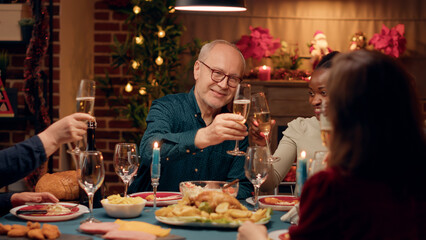 Cheerful multicultural family members toasting champagne while enjoying Christmas dinner at home. Happy close relatives clinking sparkling wine while celebrating winter feast together.