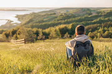 Tourist with backpack sitting on top of hill in grass field and enjoying beautiful landscape view....