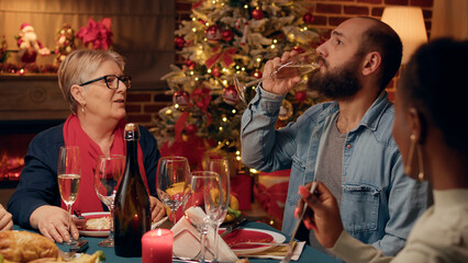 Confident man talking with mother-in-law at Christmas dinner while drinking champagne. Festive...