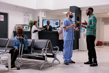 African american man talking to medical assistant in ospital reception lobby, discussing about disease diagnosis and recovery treatment. Busy waiting room with sick patients at clinic.