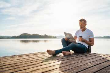 Mature man reading newspaper and having coffee at pier by lake