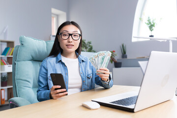 Business training about money. A young Asian business woman conducts a training session on the topic of money. He is holding a phone and a wad of bills. Sits at his desk, talks, looks into the camera.