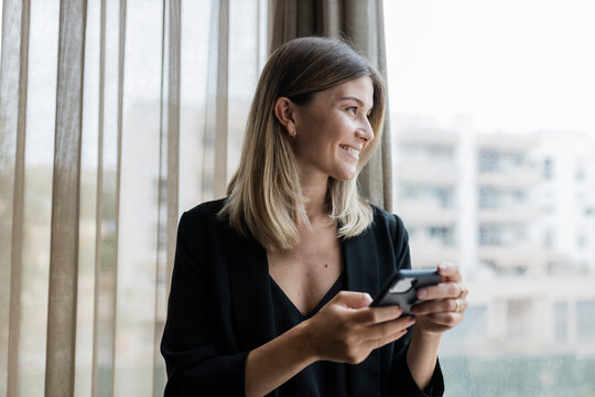 Smiling businesswoman holding mobile phone looking through window