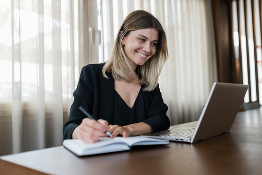 Smiling businesswoman looking at laptop screen and writing in diary on table