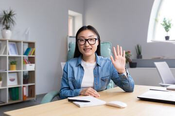 Distance Learning. A young Asian woman is a teacher, conducts online classes, lessons. He looks into the camera, explains, tells. Sitting in glasses and jeans at a table at a computer in a classroom