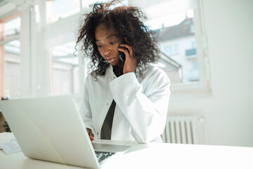 Female doctor talking on smart phone using laptop at desk