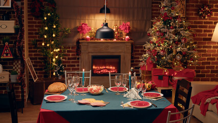 Fototapeta na wymiar Empty festive christmas dinner table inside decorated living room with holiday garlands and dinnerware. Interior of traditional and authentic season cozy setting celebrating religious event.