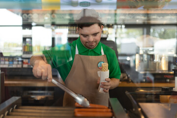 Happy waiter with Down syndrome standing by counter and prepairing hot-dog to a customer in cafe at gas station.