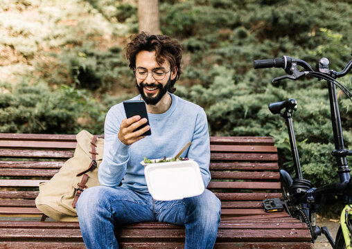 Smiling man with lunch box using phone sitting on bench
