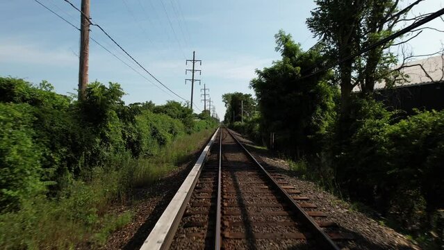 A low angle view looking straight down train tracks with green trees on either side. It runs through a Long Island neighborhood on a sunny day. The drone camera dolly in then boom up over the tracks.
