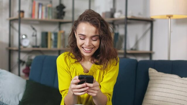 Excited young woman looking at phone screen, celebrating good news and showing yes gesture at home. Smiling attractive female receiving reward in mobile game.