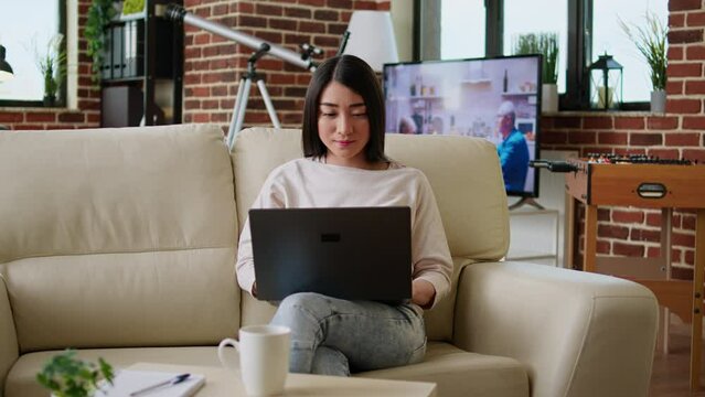 Focused young adult woman working remotely on laptop while sitting on sofa in living room at home. Attractive asian freelancer doing remote work on modern portable computer.