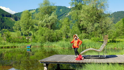 asiatische Touristin macht Selfie auf kleinem Teich im Ammergau auf Holzsteg mit Ruheliege im Grünen