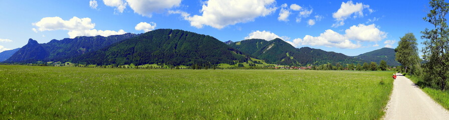 Panorama mit Wiesen, Wald und Berge im Ammergau mit Radweg unter blauem Himmel mit weißen Wolken