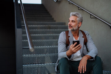 Successful happy businessman waiting in city street, sitting on stairs and calling on mobile phone, commuting concept.