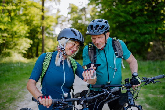 Active senior couple riding electric bicycles on road at summer park, checking their performance at smartwatch, healthy lifestyle concept.