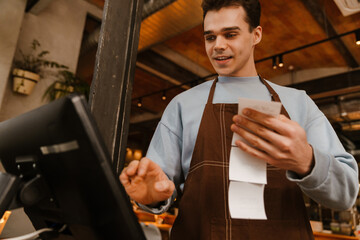 Young waiter man standing in front of computer and holding receipt