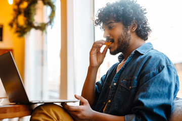 Young calm handsome indian curly man eating cake while working