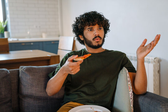 Young Indian Frowning Handsome Curly Man Sitting On Sofa Watching