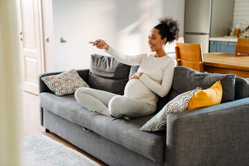 Adult pregnant smiling african woman watching tv sitting on sofa