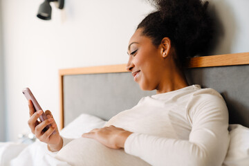 Young beautiful pregnant afro woman liying on bed looking phone