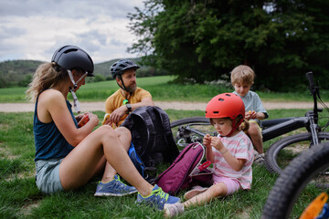 Young family with little children resting after bike ride, sitting on grass in park in summer.