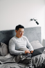 Fototapeta premium Asian man working with laptop after sleep while sitting in bed