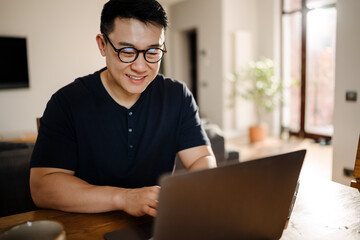 Brunette adult asian man smiling and using laptop computer at home