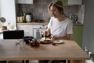 Obraz na płótnie Canvas Pensive middle-aged single woman in home wear sit alone at table in kitchen, enjoying breakfast in peace and quiet at home, mature lone lady eating healthy food in morning, spreading butter on toast