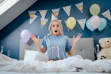 Happy woman in pajama and party cap blowing out candles on birthday cake on the bed in decorated...