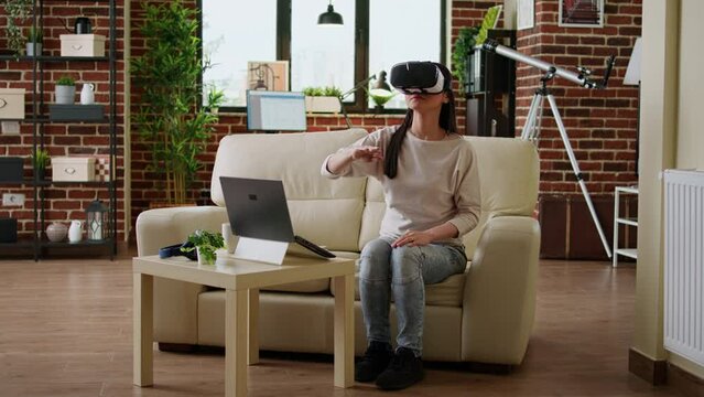 Playful woman with VR goggles playing on metaverse inside living room. Young adult person wearing futuristic virtual reality headset enjoying cyberspace while sitting on sofa at home.
