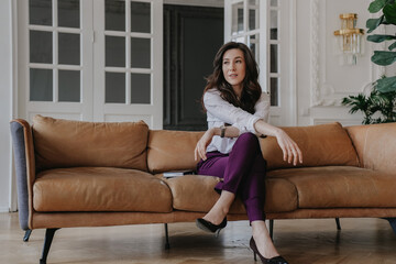 Brunette hispanic businesswoman in white shirt and violet pants sitting on leather sofa, relaxing...