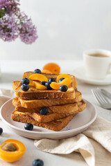Sweet french toast with berries and syrup served on plate. Copy space for text. Healthy breakfast: toasts with blueberry and apricots and a cup of coffee on white background with lilac flowers