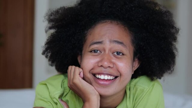 A happy black woman close-up with an afro hairstyle lies on the bed and smiles with a snow-white smile and sings songs. Portrait of afroasian woman in bedroom on bed home interior.