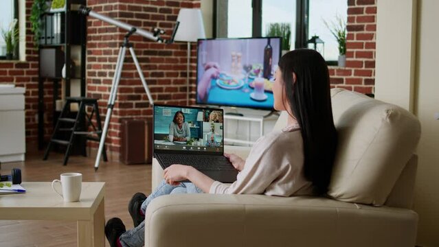 Young adult person with portable computer doing remote work while talking on online meeting. Woman sitting on couch working remotely while discussing with office manager on videocall conference