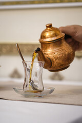 A man's hand pours tea into a Turkish cup from a copper teapot.