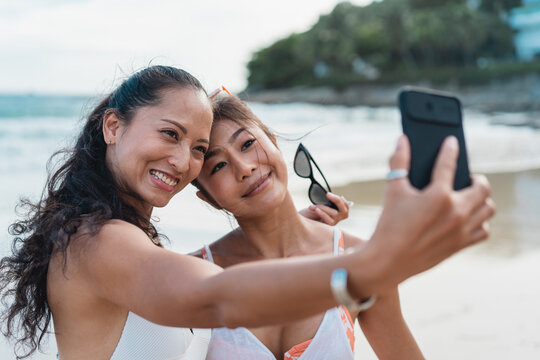 Portrait of happy smiling beautiful women taking a selfie photo with mobile phone on the beach. Pretty young Asian girls in swimwear posing and for picture together on tropical vacation