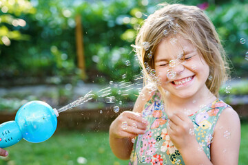 Happy little blonde preschool girl having fun with blowing soap bubble blower. Cute child playing...